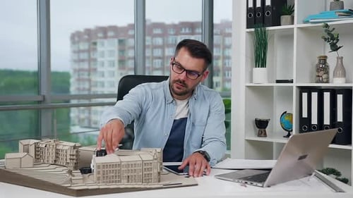 Architect in Glasses Working in Design Office with Wooden Model of New Buildings