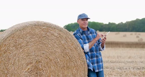 Farmer Using Tablet Beside Hay Bale in Field