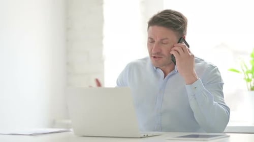 Man Talking on Smartphone at Desk with Laptop