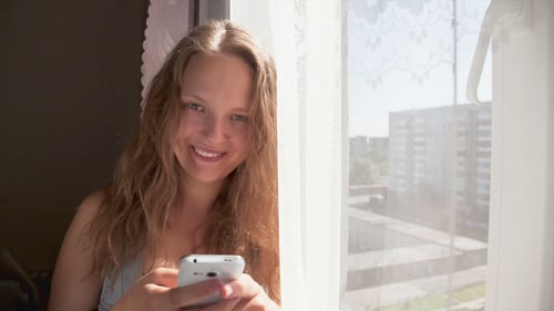 Smiling Young Woman Using Smartphone By Window