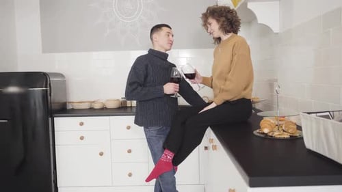 Seitenansicht einer glücklichen jungen Frau, die auf einer Tischplatte sitzt und mit einer Brille klirrt, mit kaukasischem Mann. Breit