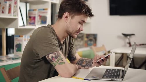 Programador atraente com tatuagens nos braços rolando algo em seu smartphone enquanto está sentado