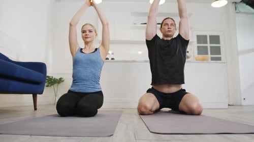Joven pareja estadounidense en ropa deportiva haciendo ejercicio de yoga en la cocina en casa por la mañana.
