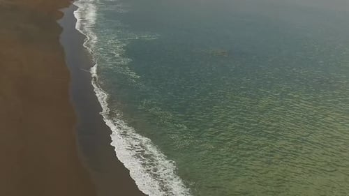 A drone flies backwards against relatively calm ocean waves drifting ashore.