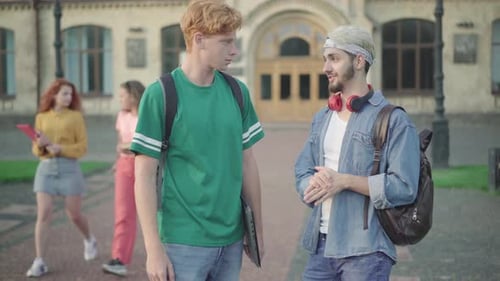 Two Young Male College Students Standing on Campus and Talking As Beautiful Women Passing at the