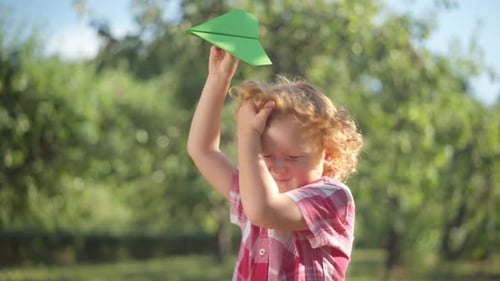 Side View Curios Redhead Little Boy with Paper Plane Standing in Sunshine Outdoors Looking Away