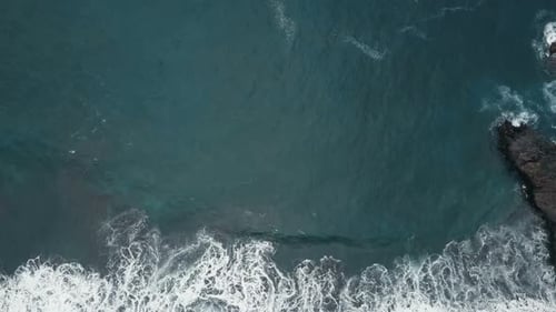 Luftaufnahme von oben von Meereswellen, die auf dunklen Felsen am schwarzen Sandstrand brechen, Kanarische Inseln, Teneriffa