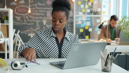 Portrait of Young African American Woman Using Laptop and Writing Data in Notebook in Office