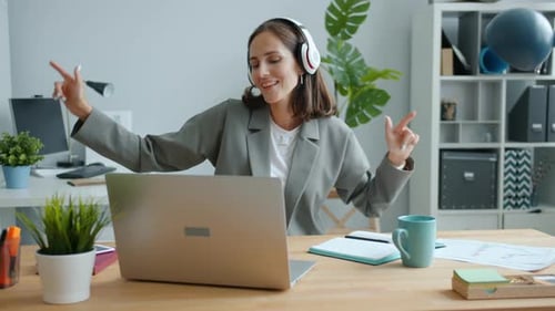 Happy Businesswoman Typing in Office Using Laptop Then Dancing Wearing Headphones