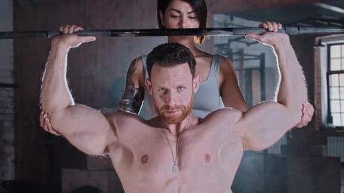 Attractive Adult Couple Training in the Gym Athletic Man Pumping His Hands Muscles with a Dumbbell