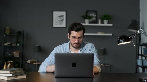 Young Business Man Sits at Table with a Laptop Working in Home Office, Typing on a Computer