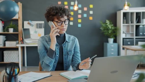 Cheerful Office Worker Talking on Mobile Phone and Looking at Laptop Screen in Modern Workplace