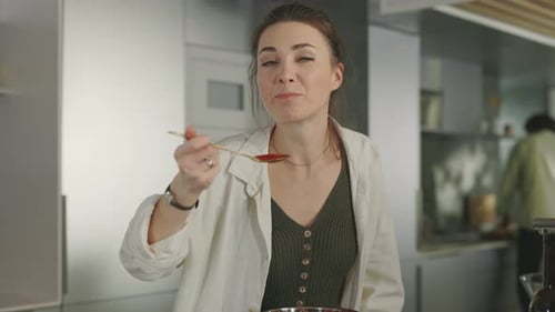 Woman Tastes Food with Spoon in Kitchen