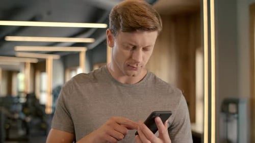 Handsome Sportsman Using Smartphone in Sport Club. Bodybuilder Standing at Gym.