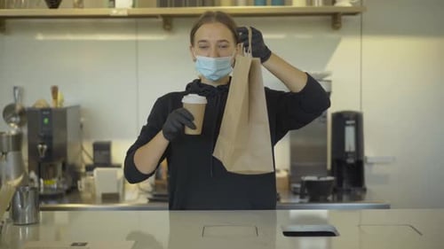 Confident Caucasian Woman in Covid-19 Face Mask Stretching Coffee Cup and Paper Bag at Camera