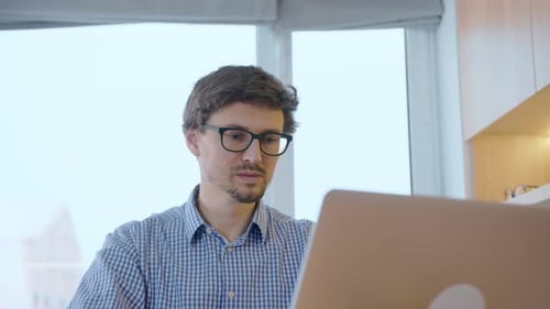 Remotely work on computer. Young man in glasses works at home at a computer, behind him is a window.