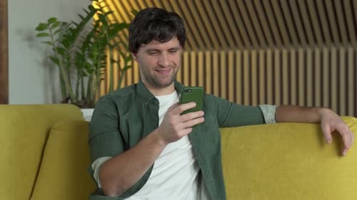 Young Man in a Green Shirt Uses a Smartphone at Home on a Yellow Sofa