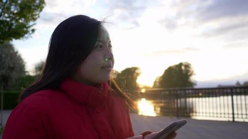 Close up Asian woman using smartphone wearing red coat sitting on the chair at the park