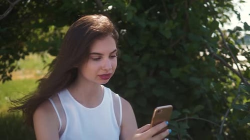 Beautiful Cheerful Girl with a Smartphone Sitting in a Park on a Bench on a Sunny Day Teenager