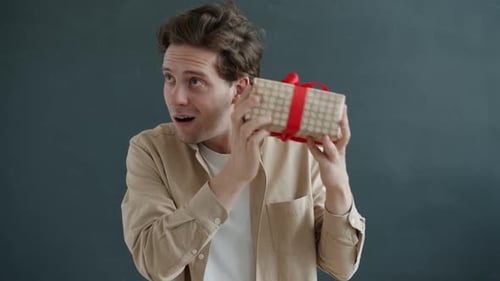 Slow Motion Portrait of Cheerful Guy Shaking Gift Box Enjoying Present and Surprise