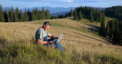 A Man Sits on a Beautiful Meadow in the Mountains Works on a Laptop