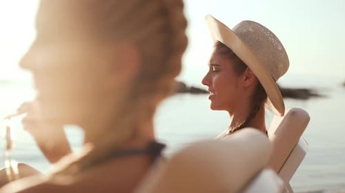Two Beautiful Girls Relaxing at the Sea Side Sitting in Chaise Lounges