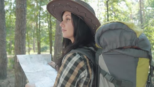 Charming Young Caucasian Female Tourist with Map Looking Around in Sunny Forest and Walking Away