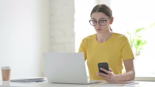 Woman Browsing Internet on Smartphone while using Laptop in Office