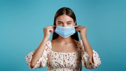 Woman Puts On Surgical Mask Against Blue Background
