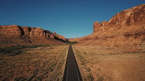 Aerial Shot of Car Driving Along Straight American Desert Highway Road Among Atmospheric Rocky
