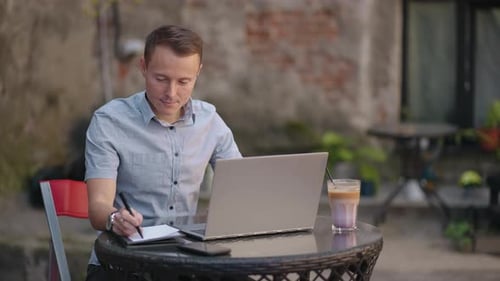 A Man Sitting in a Street Cafe Works Remotely Writing a Pen in a Notebook