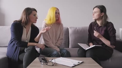 Three women talk while sitting on gray couch