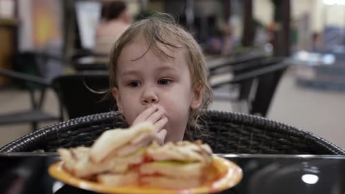 A Small Blonde Girlchild Eating a Large Sandwich in Parts Sitting at a Table in the Cafe of the