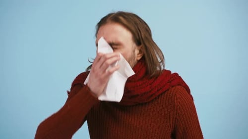 Young Guy is Sneezing Into White Napkin Looking Unhappy and Sick While Posing Against Blue Studio