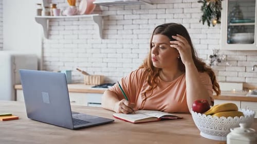 Konzentrierte Studentinnen in Übergröße schauen sich Webinar auf dem Laptop an und machen Notizen in der Küche