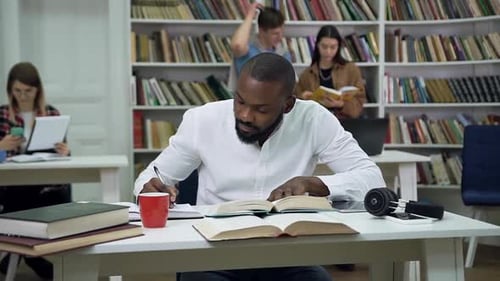 Bärtiger junger afroamerikanischer Student sitzt in der Bibliothek und arbeitet über die Aufgabe zu Hause