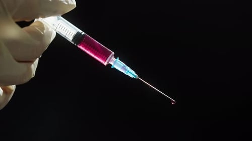 Mão em luvas médicas de proteção segurando seringa com solução vermelha em fundo preto