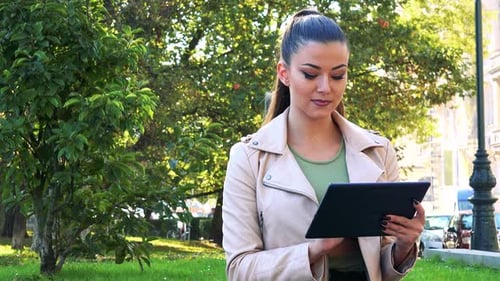 Young Beautiful Woman Works on Tablet in the City - Park in the Background