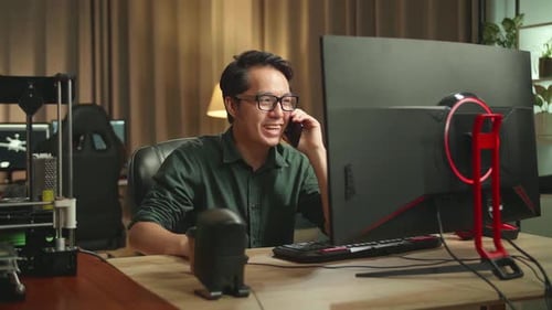 Asian Man Talking On Mobile Phone While Works On Personal Computer And 3D Printer In Home Office