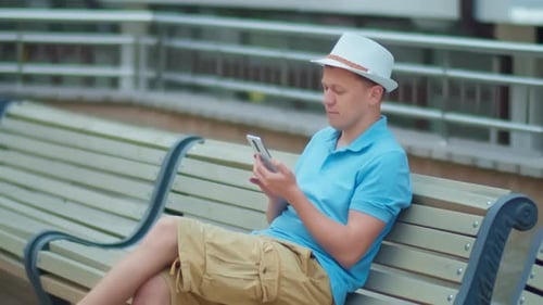 Young man in hat sits on a city bench with a mobile phone in his hands, camera tracking