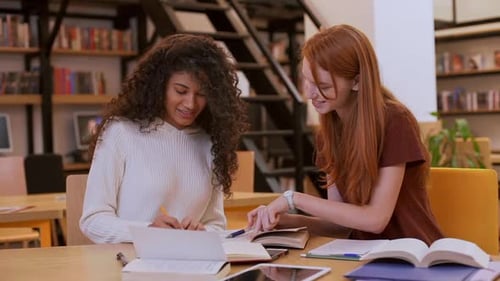 Two Teenage Girls Sitting at Library While Boy in Shirt and Glasses Coming to Them Discussing Book