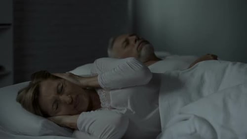 Homme ronflant dans son lit, femme à côté de lui pressant les mains contre les oreilles, sommeil troublé