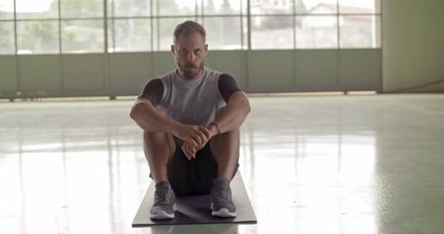 Muscular Man Sits On Yoga Mat In Bright Gym