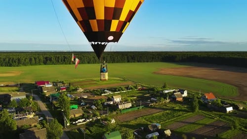 Two aeronauts in gondola fly balloon. A yellow balloon over village at sunset.