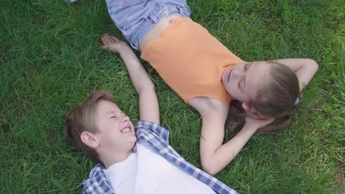 Two Funny Kids Lying on the Grass in the Park Smiling To Each Other. The Girl Taking Hand of the Boy