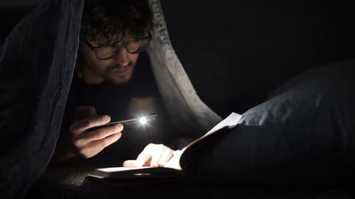Mann liest nachts ein Buch mit einer Taschenlampe unter der Decke