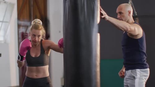 Caucasian female boxer wearing boxing gloves training with punching bag with male trainer at the gym
