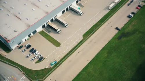 Aerial following shot of semi-trailer trucks travelling along a warehouse on the parking lot in the