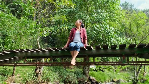 A beautiful Asian woman sits on a wooden bridge across a stream in a mountain forest and enjoys.
