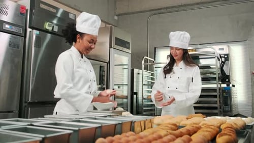 Female chefs in uniforms prepare to bake bread and pastry in stainless kitchen.